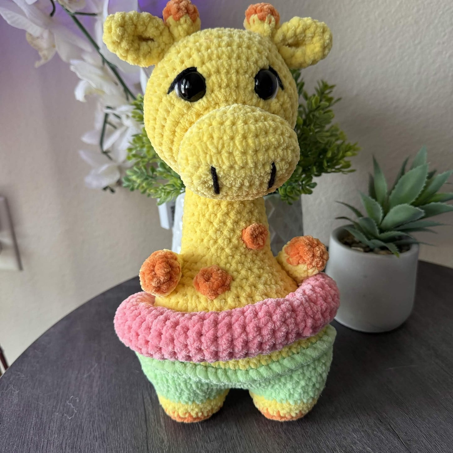 Crocheted giraffe toy with a pink floatie on a table with plants in the background
