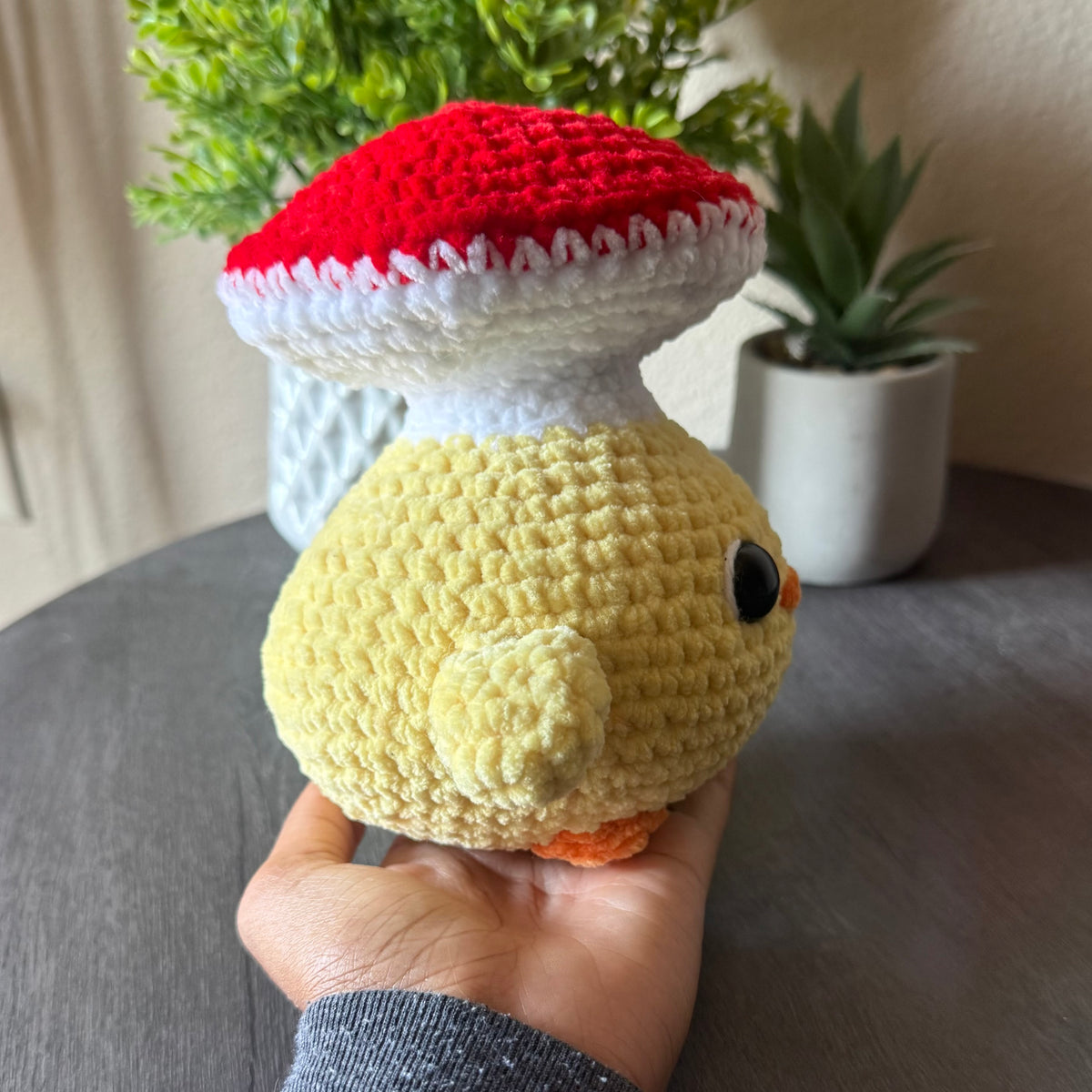 Hand holding a small crochet mushroom toy with a red cap and white stem against a neutral background.