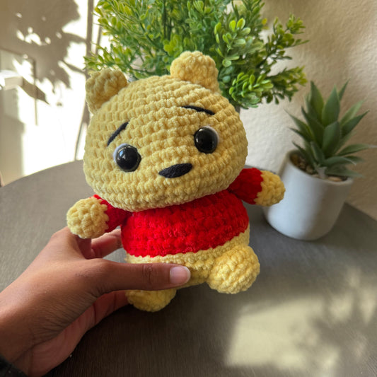 Hand holding a small crochet bear toy with a red shirt in front of indoor plants.