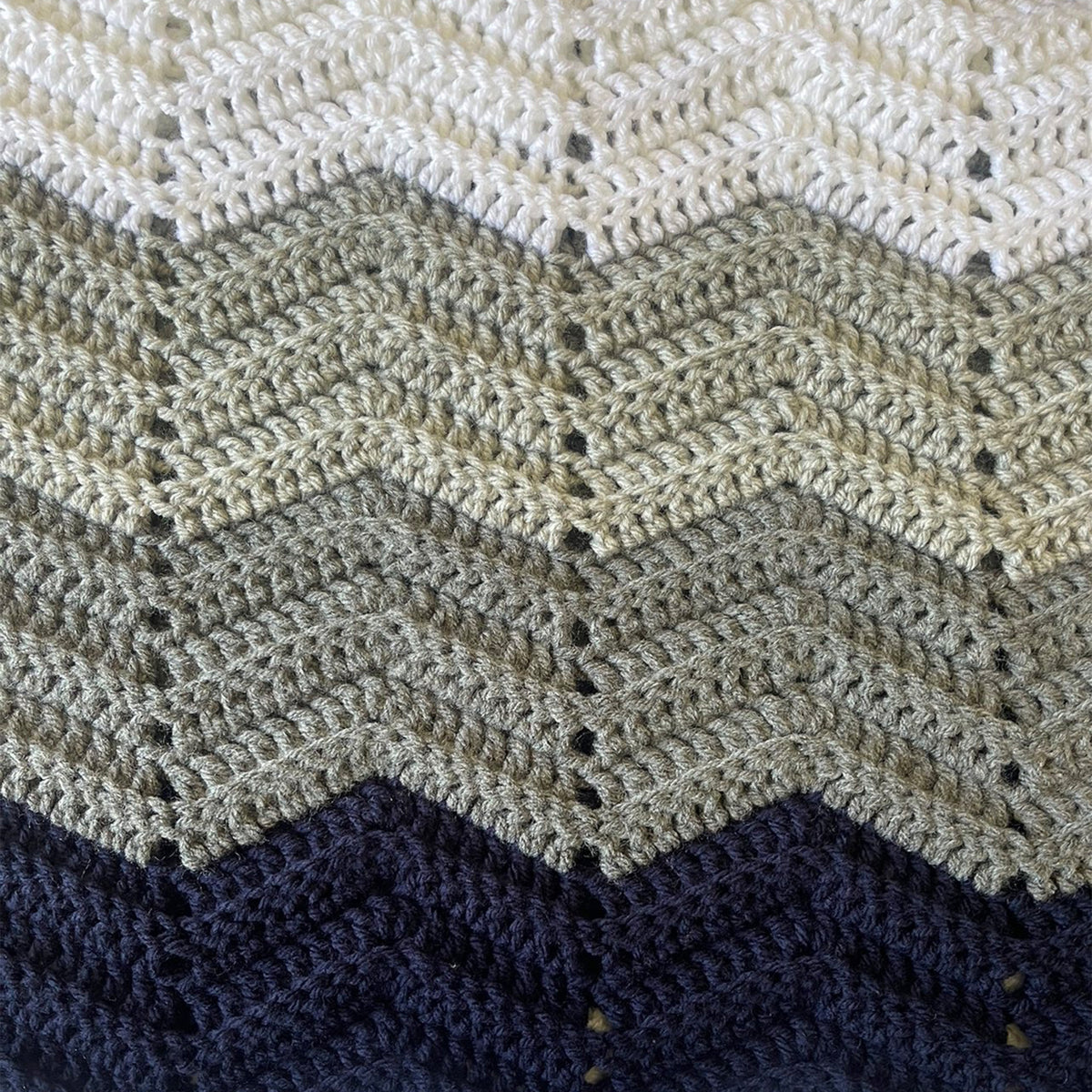 Close-up of a crocheted textile with a chevron pattern in three horizontal color bands: off-white at the top, warm beige in the middle, and dark navy at the bottom.