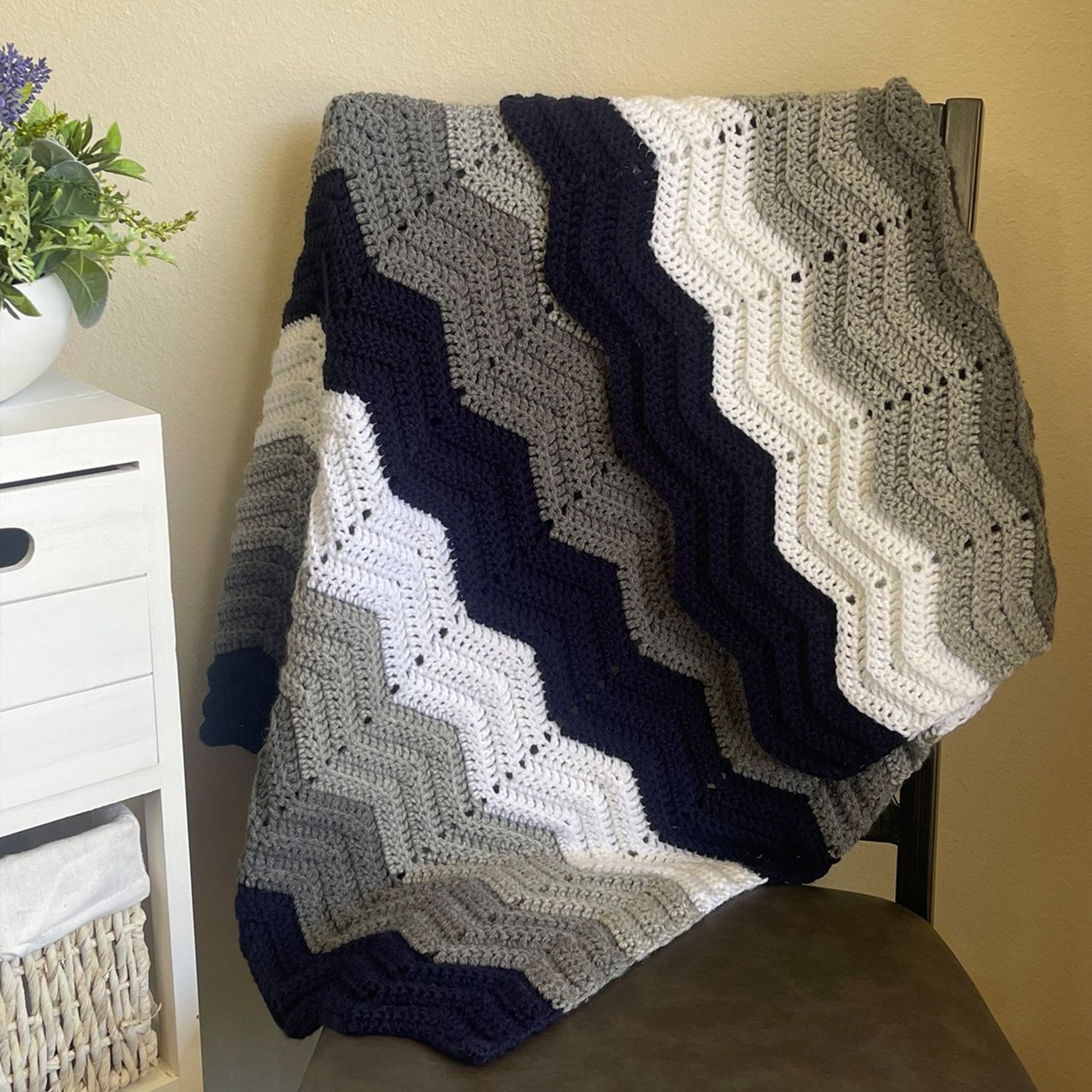 A crocheted chevron blanket with alternating zigzag stripes in navy, gray, white, and light beige, draped over a chair in a home setting.