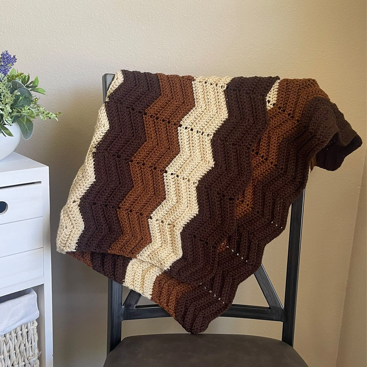 A crocheted chevron blanket in alternating stripes of dark brown, medium brown, rust orange, and cream is draped over a dark wooden chair in a home setting.