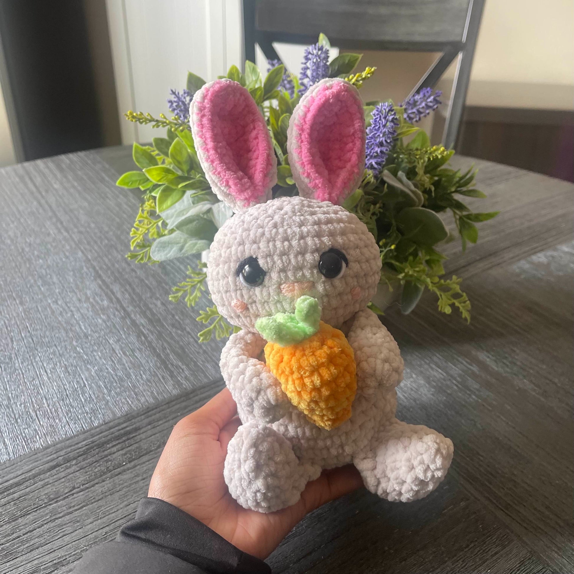 Hand holding a small crochet bunny with pink ears and a carrot, with a plant in the background.