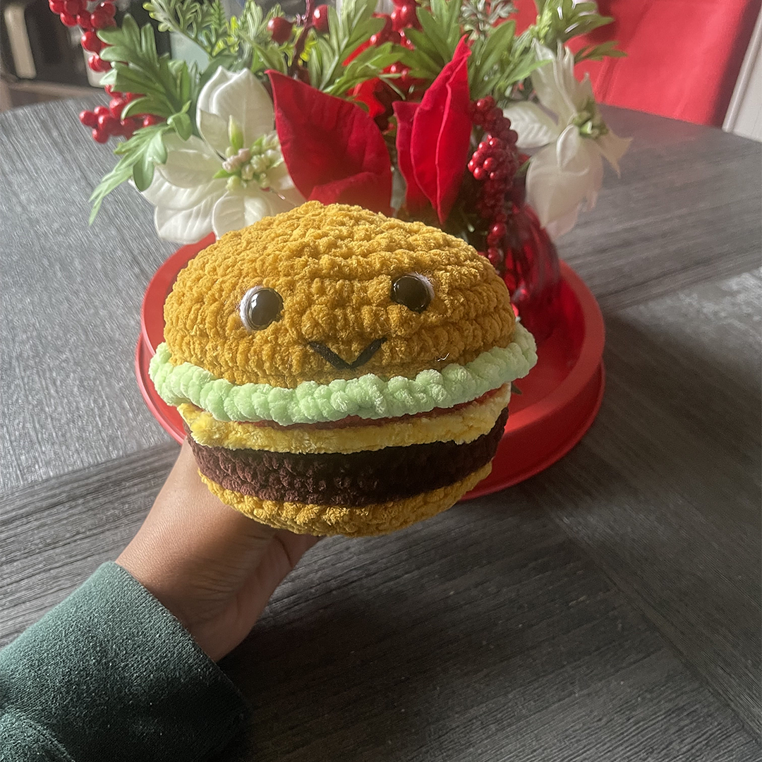 Hand holding a crocheted hamburger toy with a decorative flower arrangement in the background.