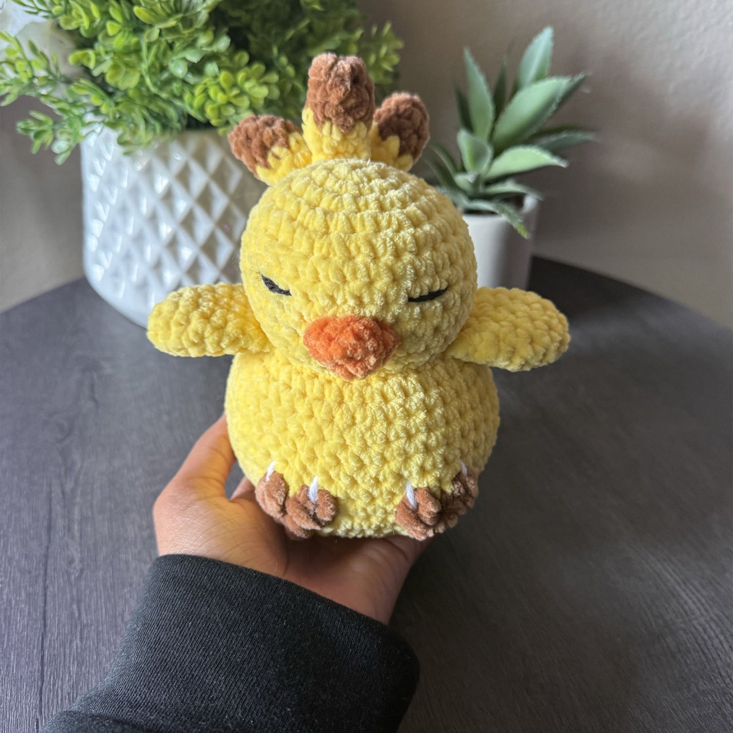 A yellow crocheted chick plush toy being held in a person's hand, with a dark gray tabletop and potted green plants in the background.