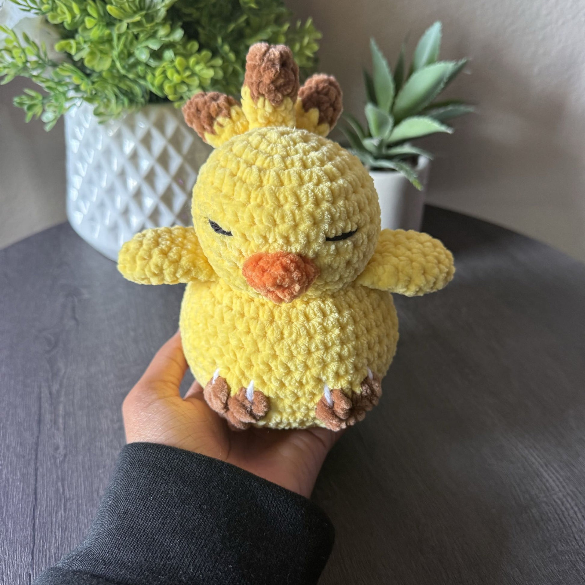 A yellow crocheted chick plush toy being held in a person's hand, with a dark gray tabletop and potted green plants in the background.