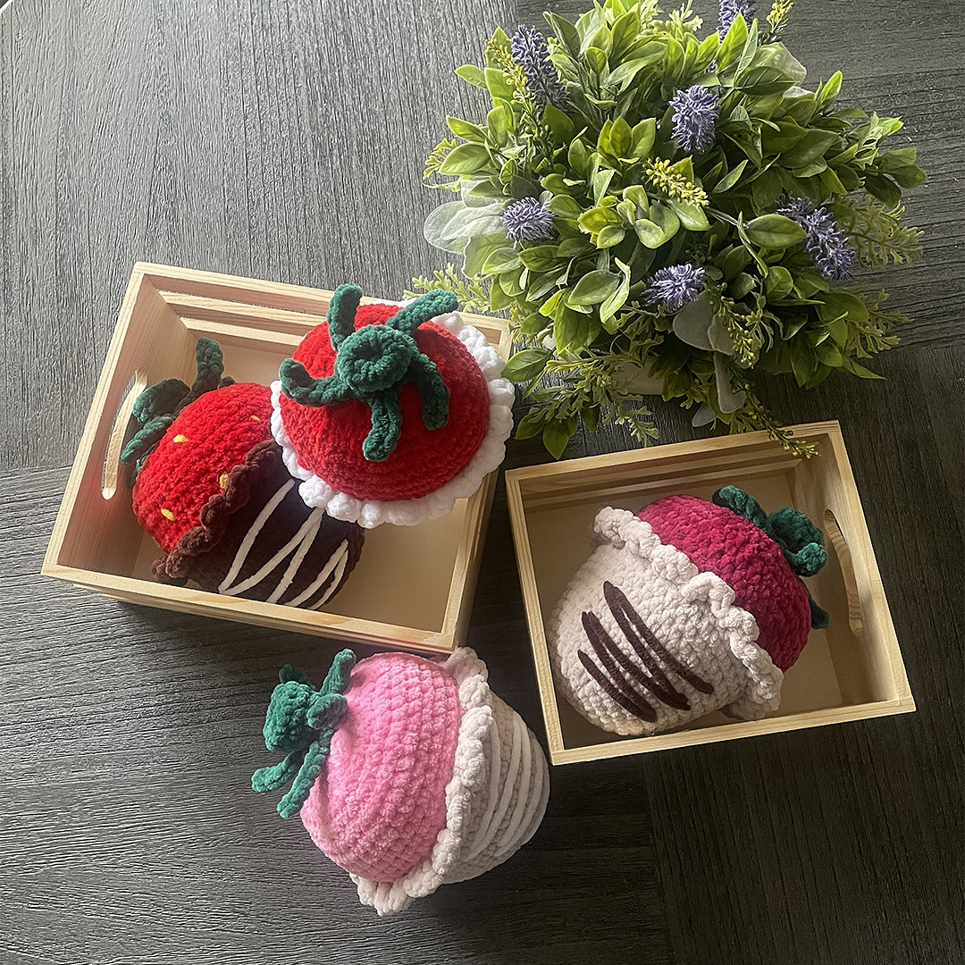 Crocheted strawberry plushies in wooden boxes with a bouquet of flowers on a wooden surface.