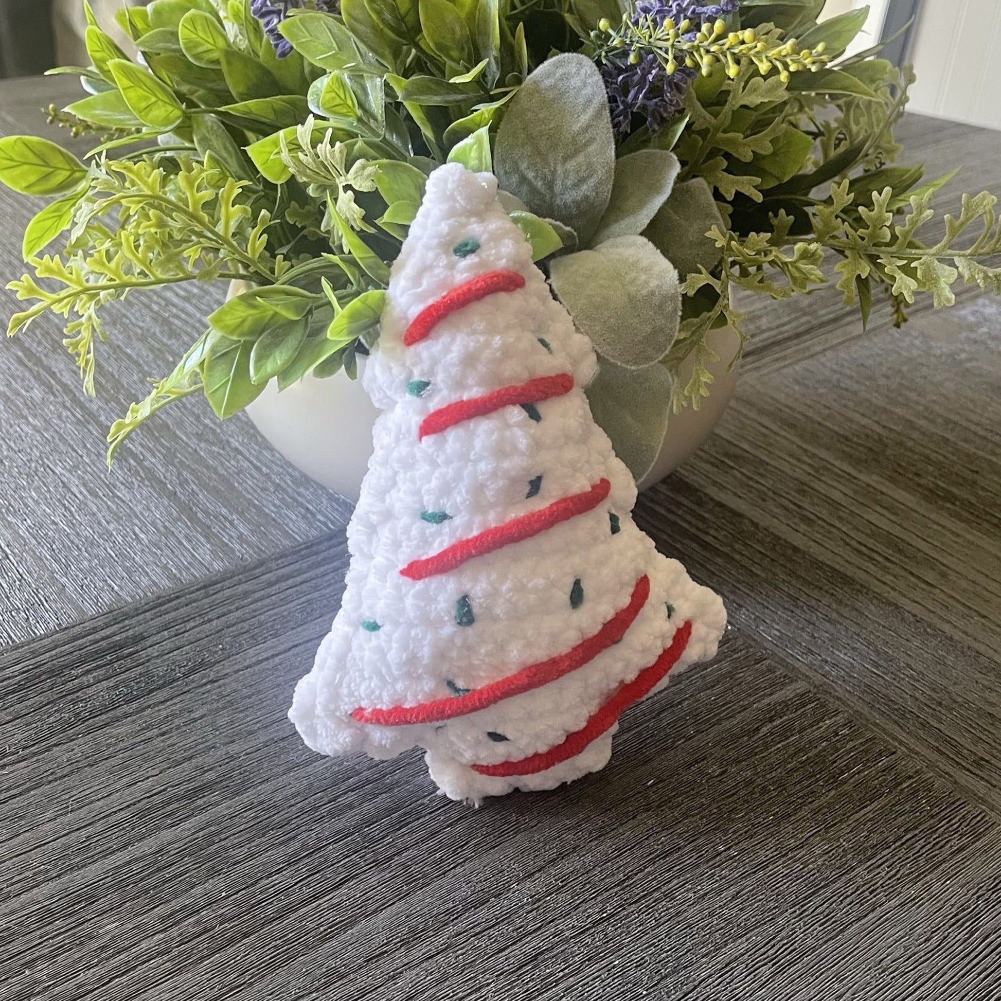 A small white plush Christmas tree ornament with red stitched stripes and tiny green accents, standing on a gray wood surface in front of a potted plant.