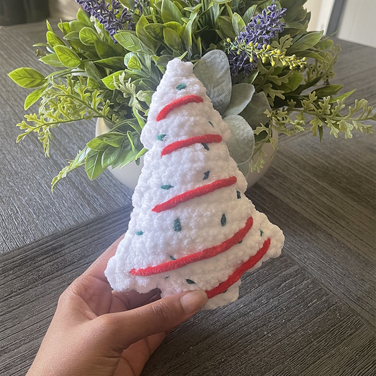 Hand holding a small, white, textured Christmas tree ornament with red stripes in front of a plant.