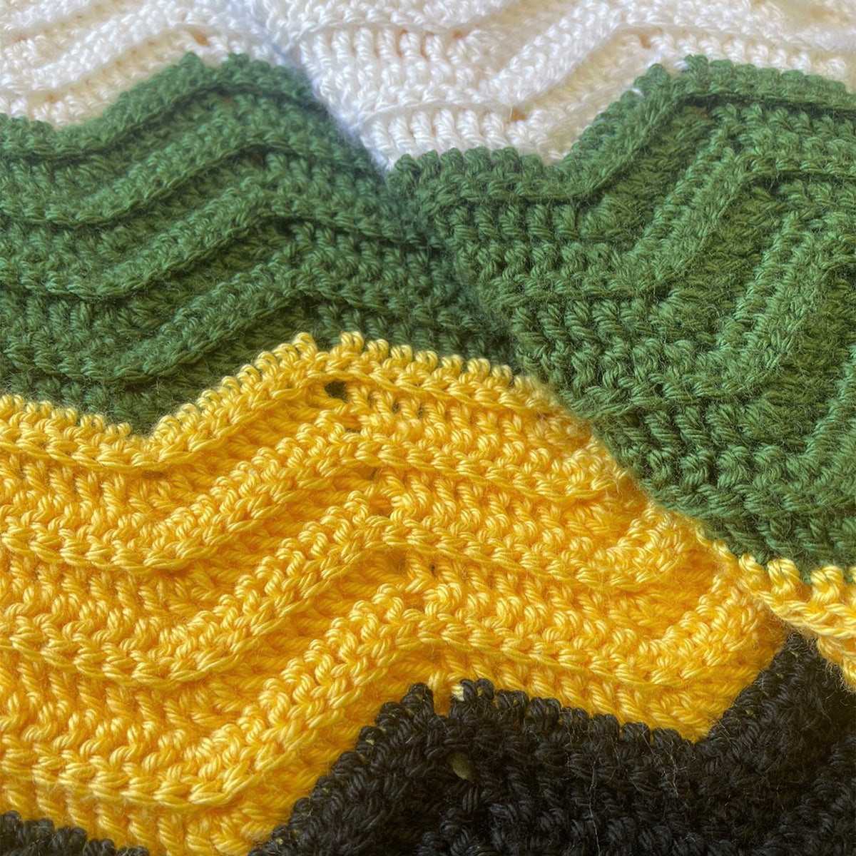 Close-up of a crocheted textile with a chevron pattern in white, olive green, mustard yellow, and dark brown.