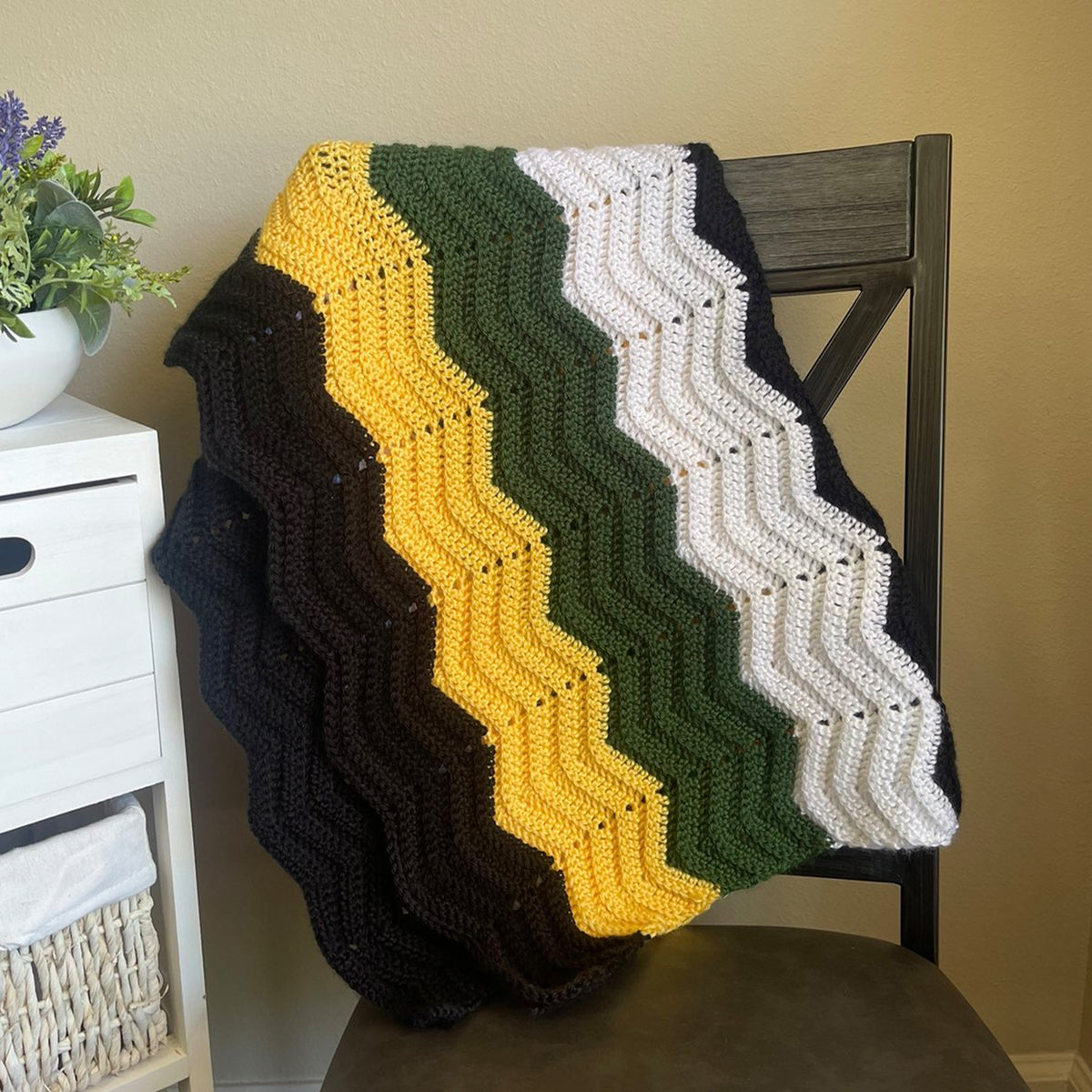 A handmade crocheted blanket with chevron stripes in black, dark brown, mustard yellow, olive green, and white, draped over a dark wooden chair in a home setting.