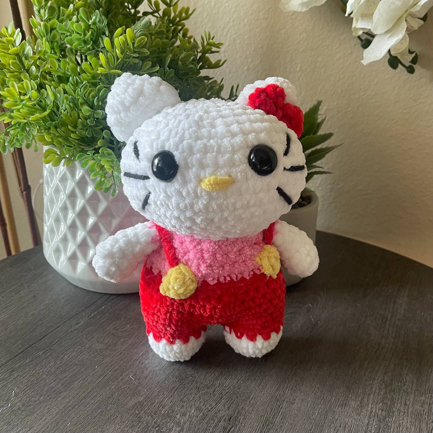 Crocheted Hello Kitty toy in red overalls on a wooden surface with plants in the background
