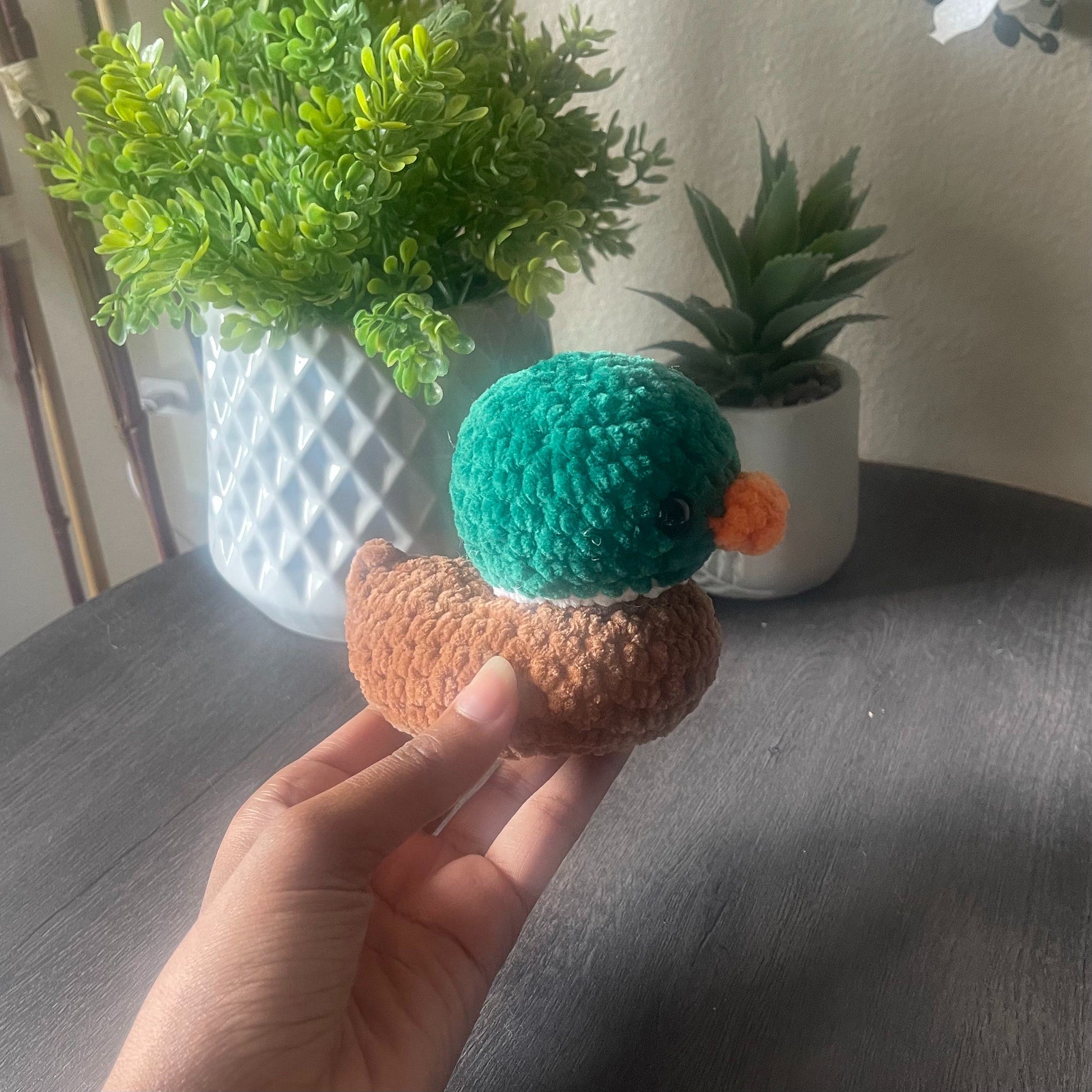 Hand holding a small crocheted mallard duck with plants in the background