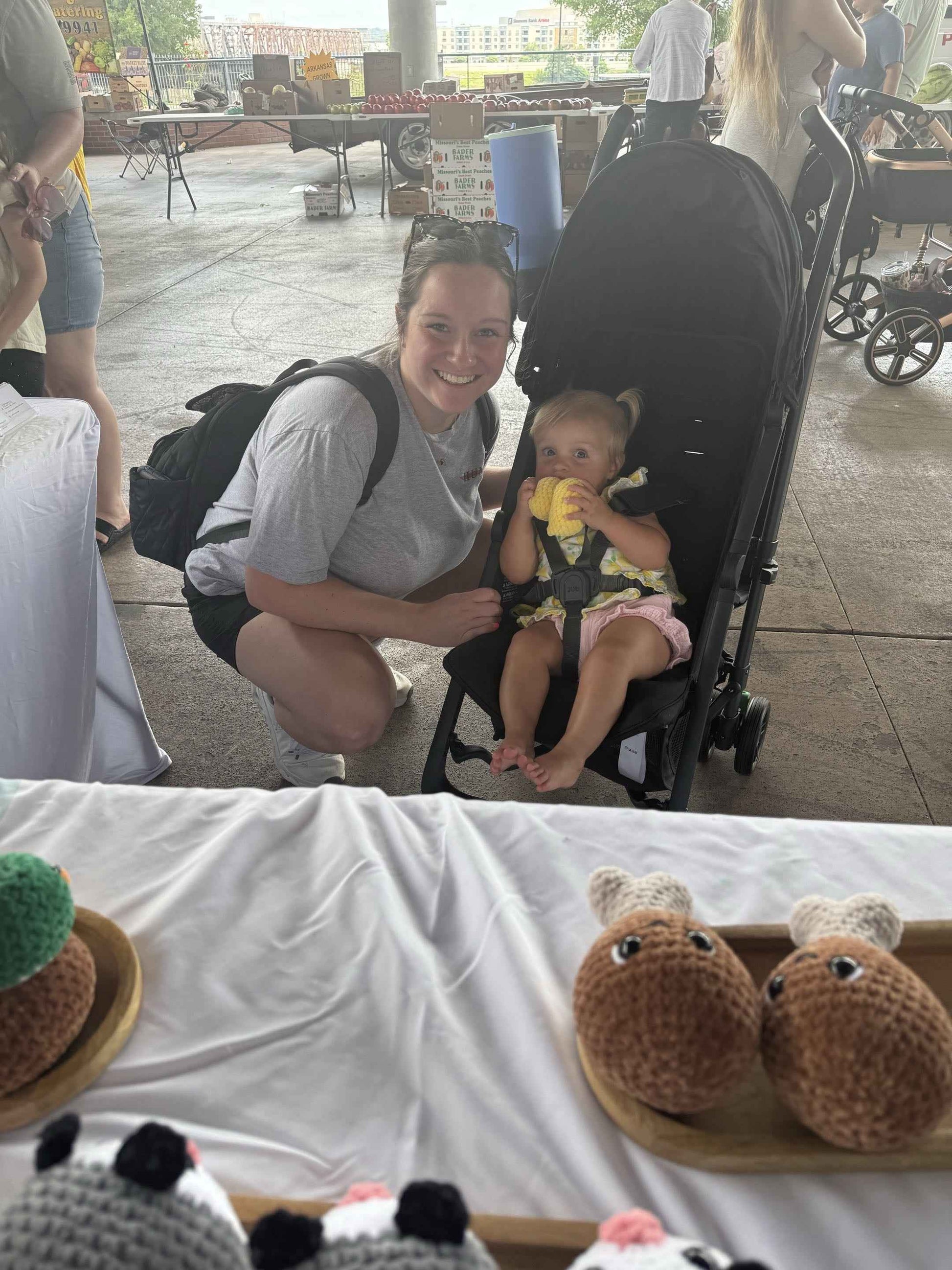 Woman with a baby in a stroller at an outdoor event with various crochet items on display.