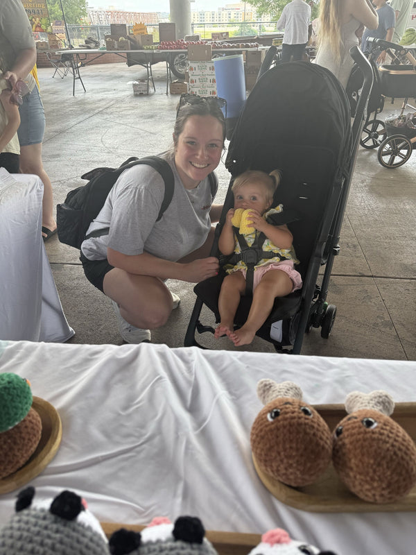 Woman with a baby in a stroller at an outdoor event with various crochet items on display.