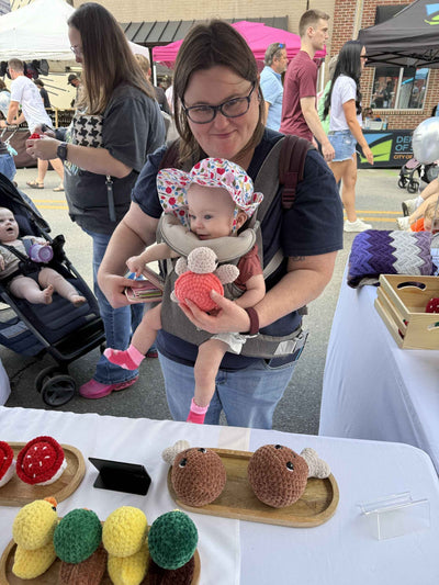 Woman holding a baby at an outdoor event with a table displaying yarn crafts.