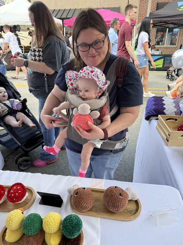 Woman holding a baby at an outdoor event with a table displaying yarn crafts.