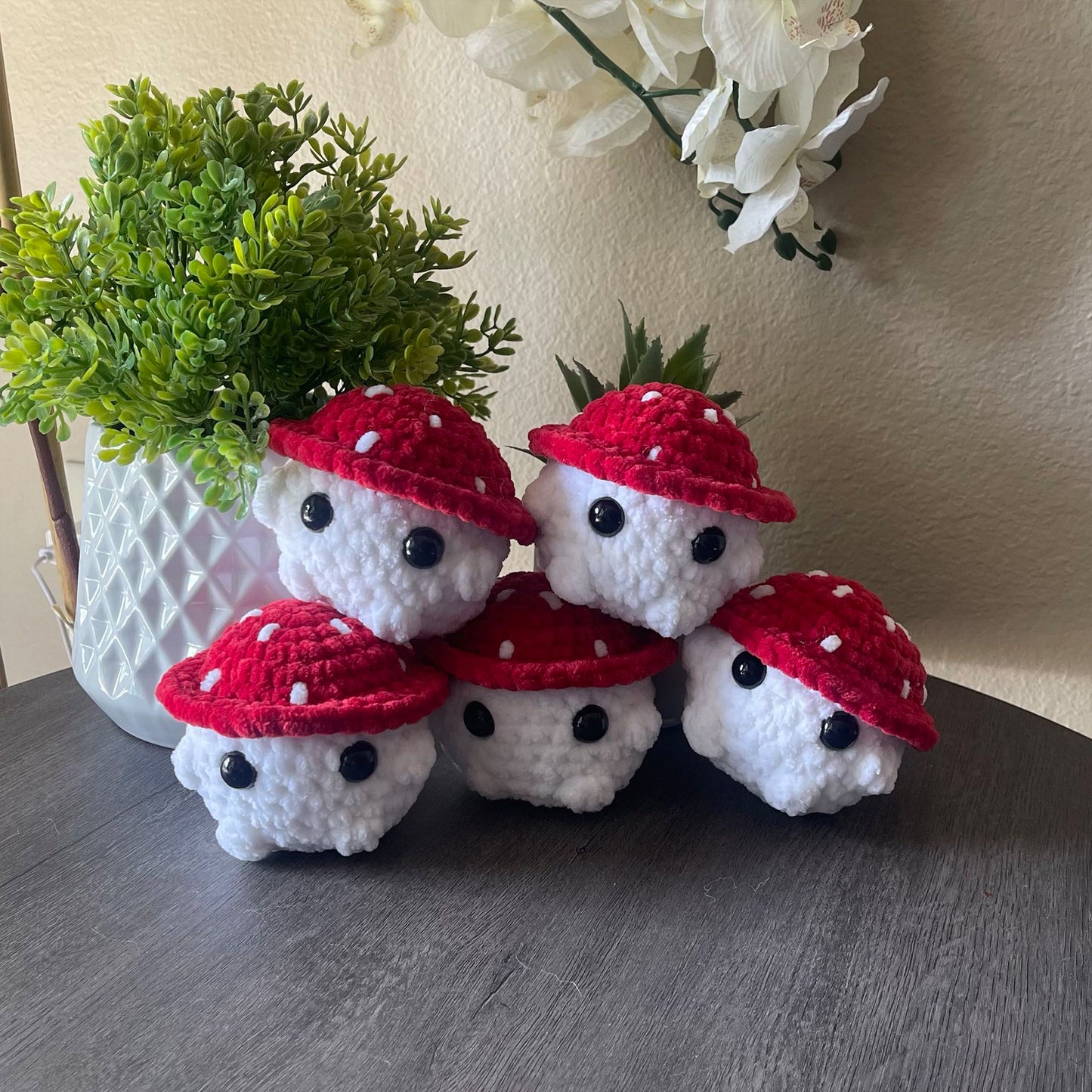 Five small crocheted plush mushroom characters with red caps and white bodies are arranged on a dark tabletop indoors, with a potted green plant and white wall flowers in the background.