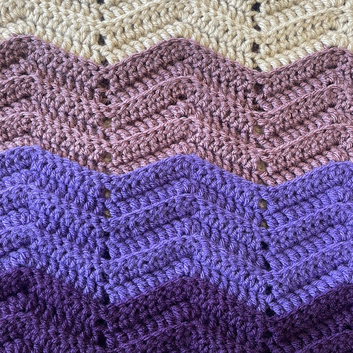 Close-up of a crocheted textile with wavy color-blocked stripes in cream, dusty pink, lavender, and deep purple, showing the textured stitch pattern.