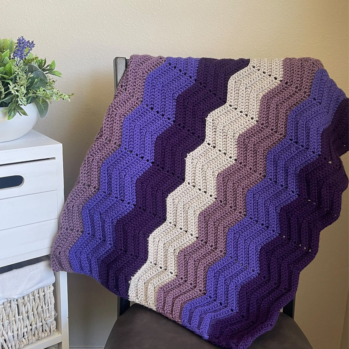 A crocheted throw blanket with chevron stripes in purple, mauve, and cream, draped over a dark chair in a home setting next to a white side table with a plant.