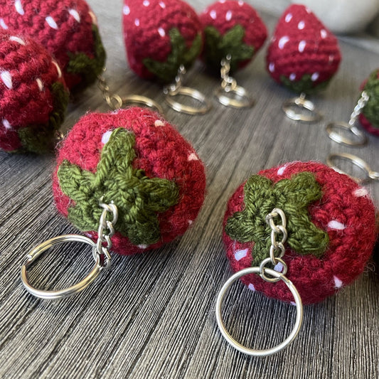 A close-up of several crocheted strawberry keychains arranged on a gray wood surface. The keychains feature red and green yarn with small white yarn accents representing seeds, attached to silver metal key rings.