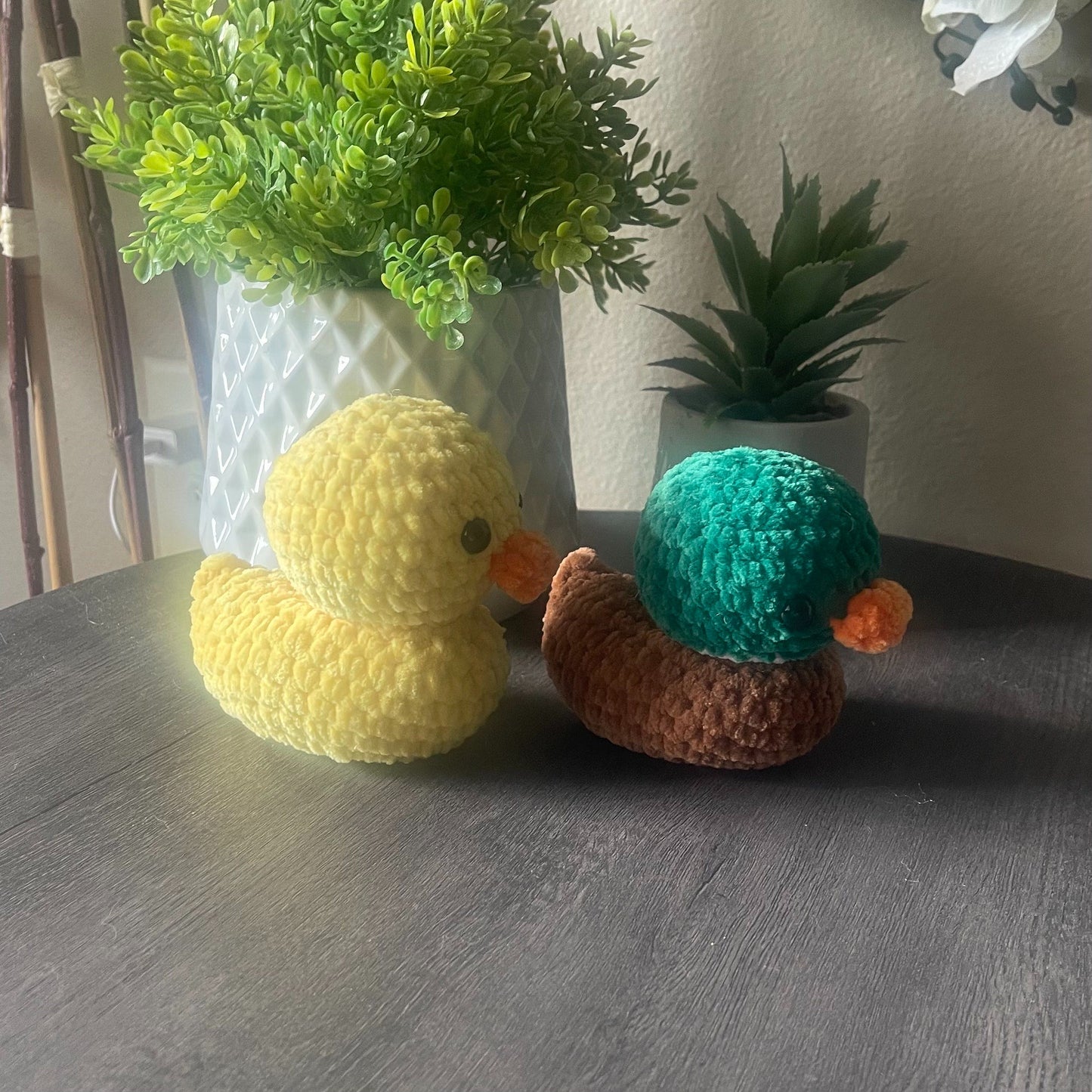 Two crocheted toys, a yellow duck and a mallard duck on a wooden surface with plants in the background