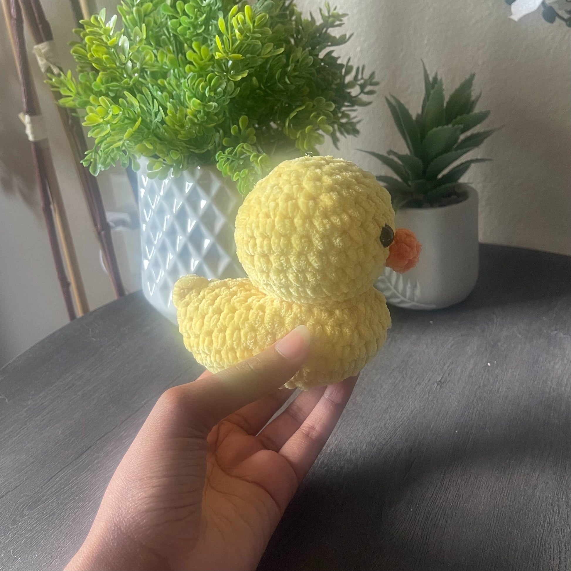 Hand holding a yellow crocheted duck toy with plants in the background